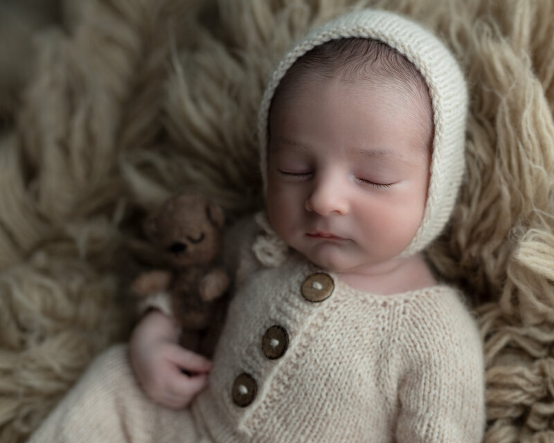Newborn boy in soft cream knits, peacefully holding a teddy on a textured flokati rug—styled in rustic tones.