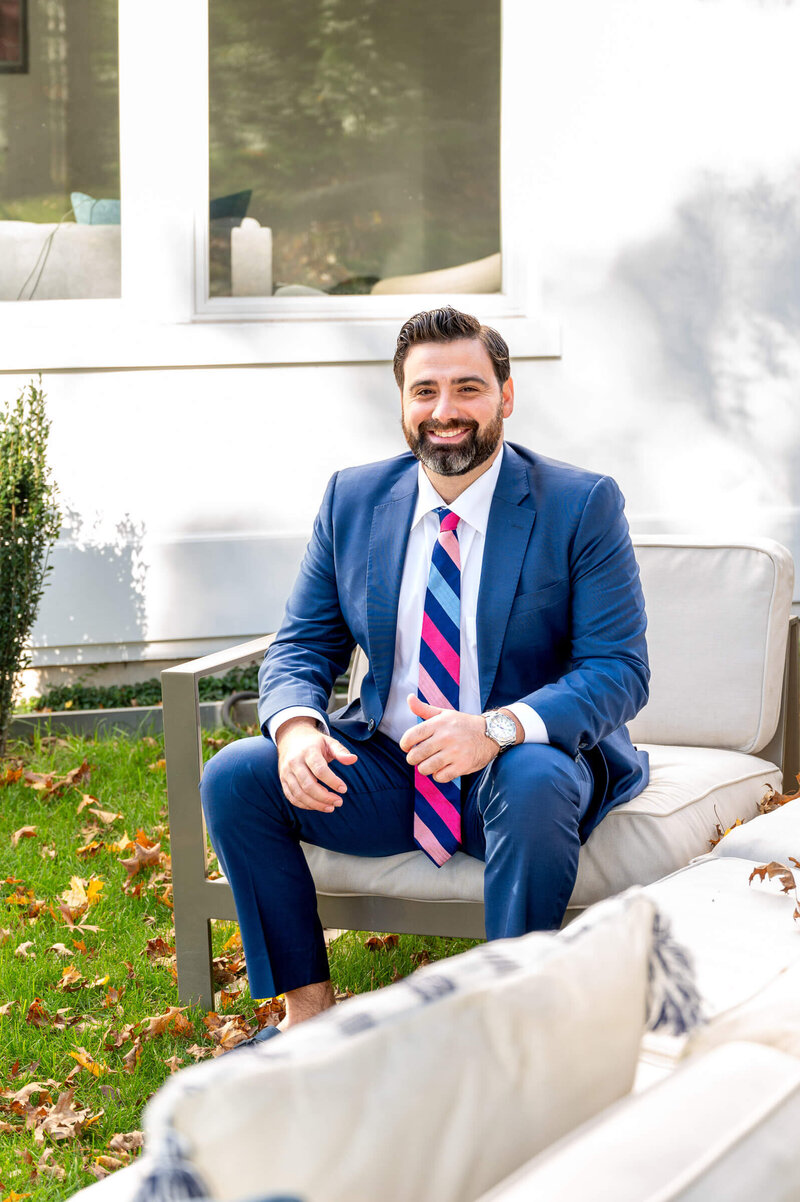 Young man in a blue suit and a pink and blue colorful striped tie, sitting outside on white patio furniture smiling  on a beautiful fall day with leaves on the ground.