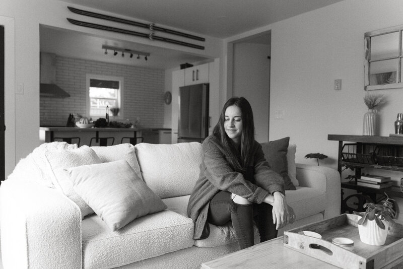 Woman with brown hair and a dark jacket and jeans sitting on a couch with a soft smile and glancing on the floor
