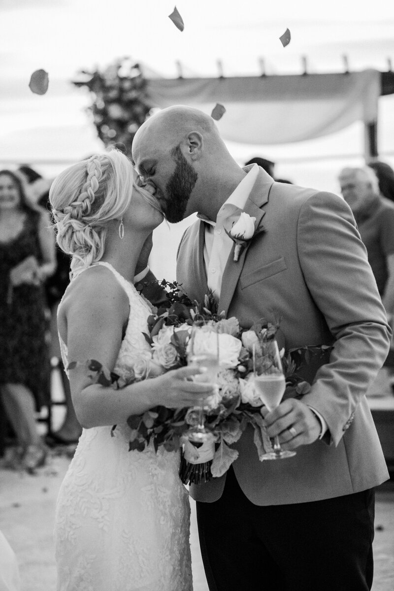 A bride and groom kiss after their wedding ceremony. The bride is holding a bouquet of flowers and a glass of champagne, while the groom, dressed in a light-colored suit, holds her close. Guests and flower petals can be seen in the background.