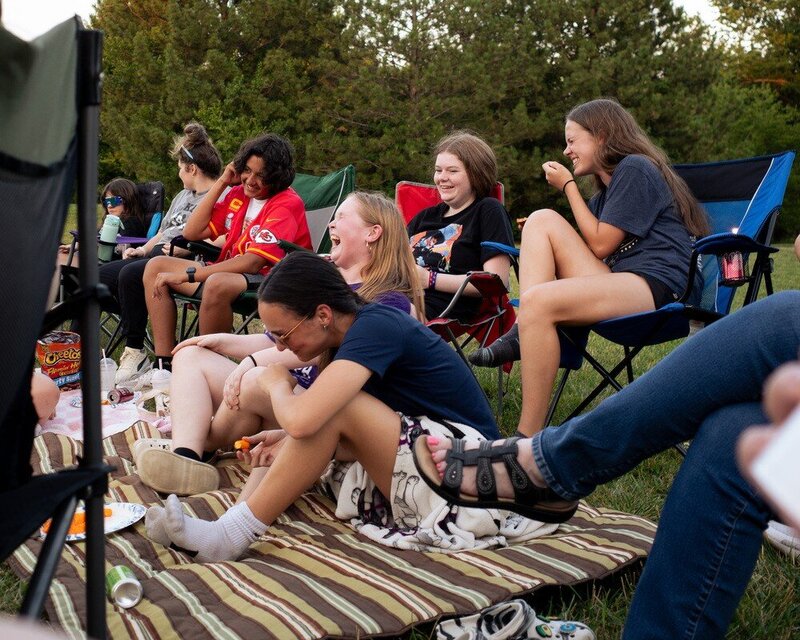 Group of people laughing four sitting in folding lawn chairs and two sitting on the ground on a blanket 