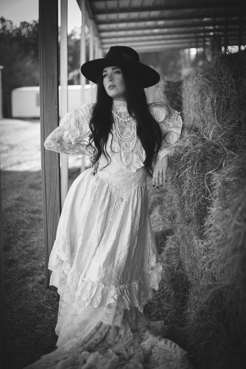 A stunning black and white photo captured by a talented destination wedding photographer. The image showcases an elegant woman in a beautiful white dress leaning gracefully against rustic hay bales.