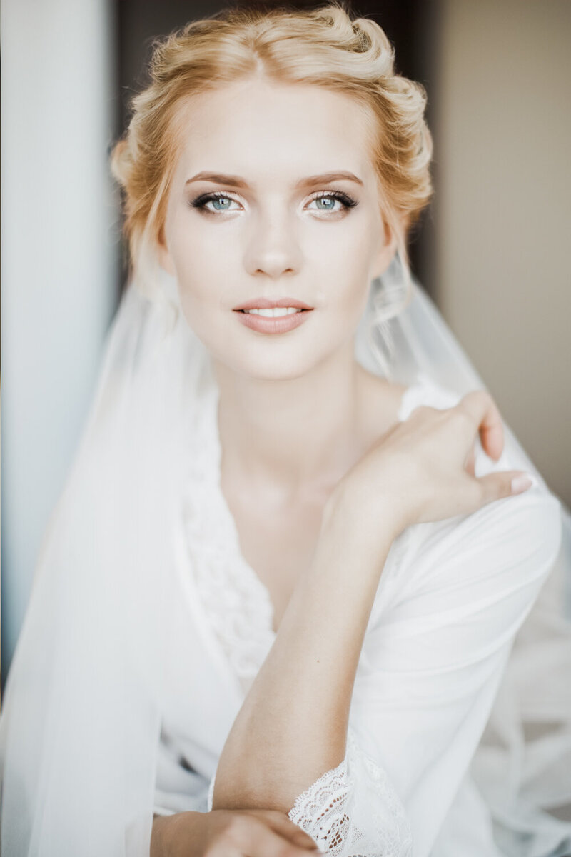 A bride poses in a white robe while getting ready for her wedding day.