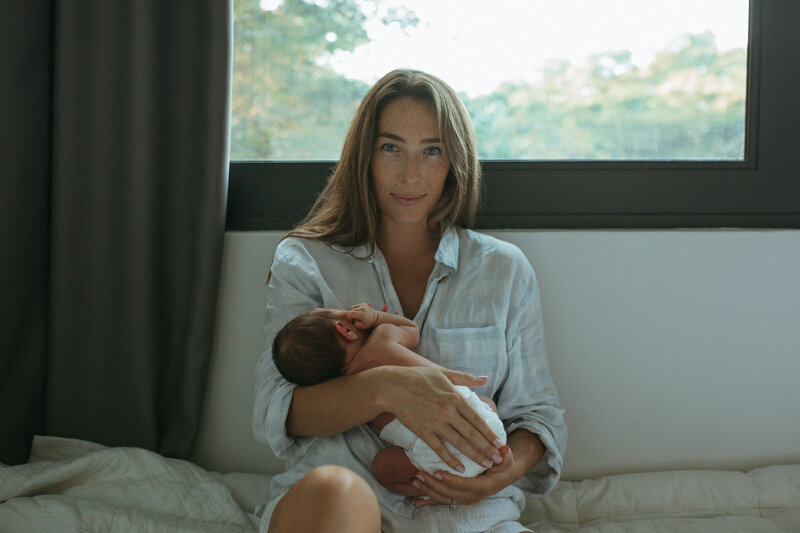 A mother sits in front of a window clutching her newborn and gazes at the camera