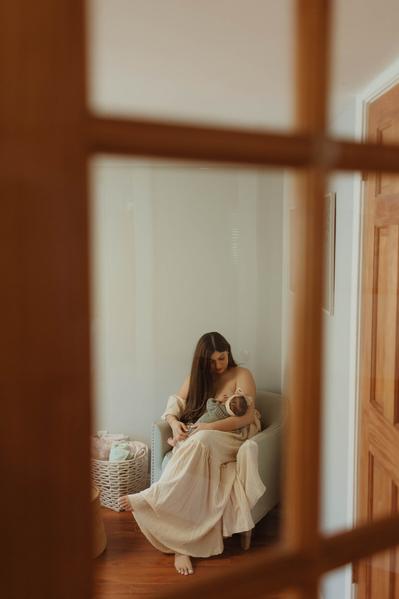 Candid moment of mother breastfeeding her newborn in nursery, captured through glass door during relaxed newborn session in Los Anegeles 
