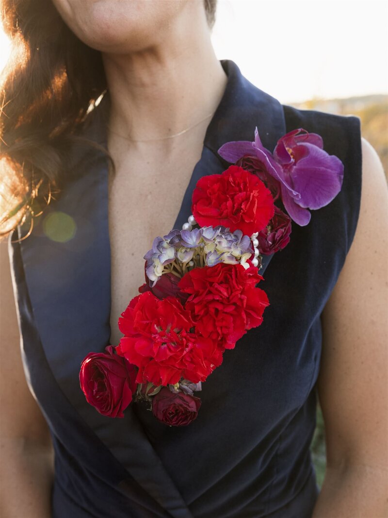 Woman in a navy blue velvet sleeveless jumpsuit with a red carnation, red rose, purple hydrangea, purple lily arrangement along her left lapel