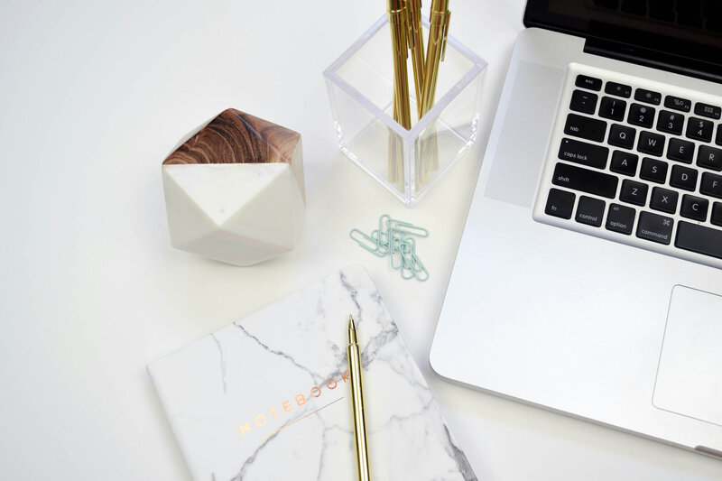 A marble-patterned notebook with a gold pen rests on a white surface beside a laptop. Nearby, a geometric container, clear pen holder with gold pens, and scattered paper clips evoke the meticulous organization of personalized travel planning.
