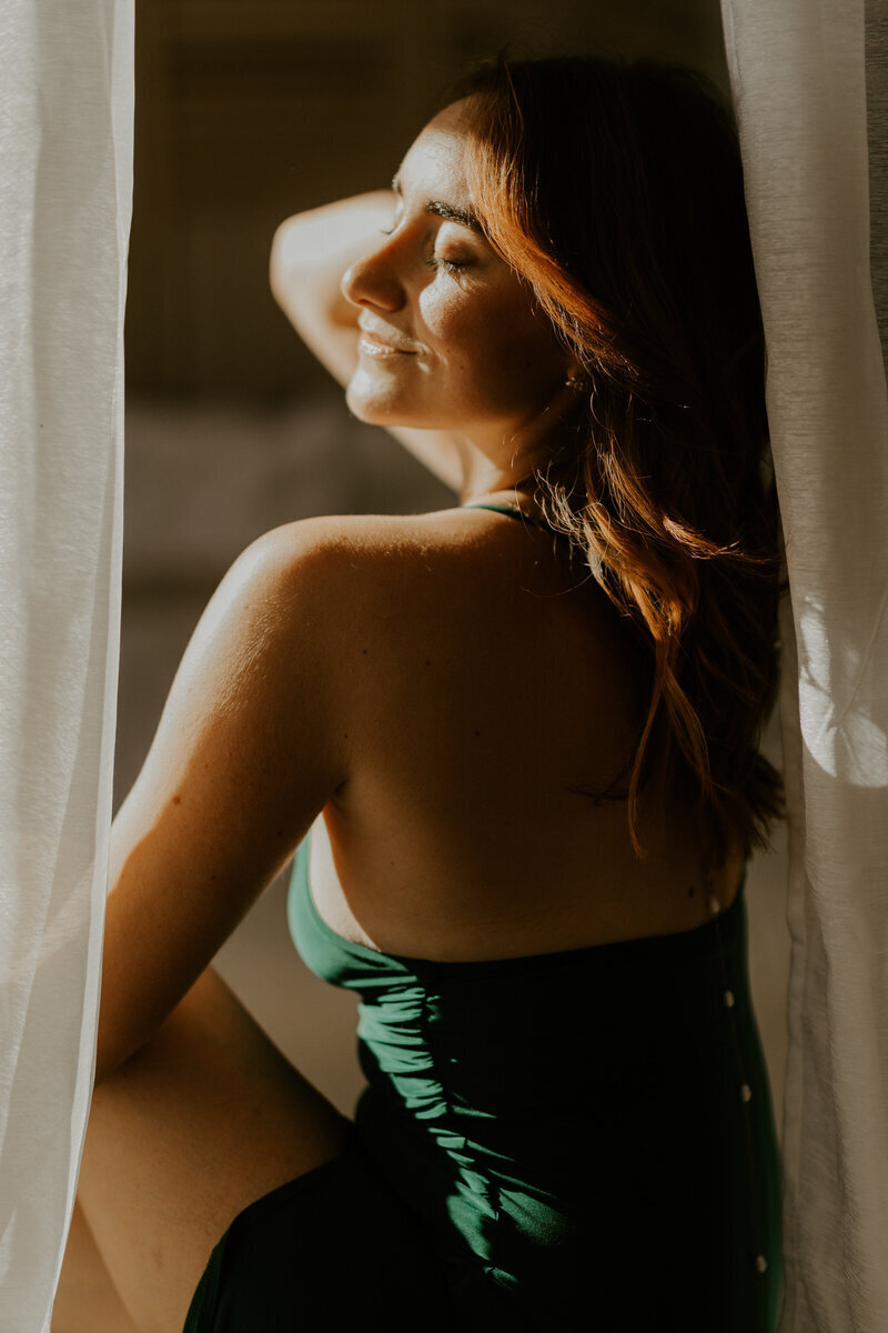 Femme rousse souriante, de dos, avec haut vert, visage tourné vers une lumière douce, derrière un voilage, à mon studio photo en Vendée.