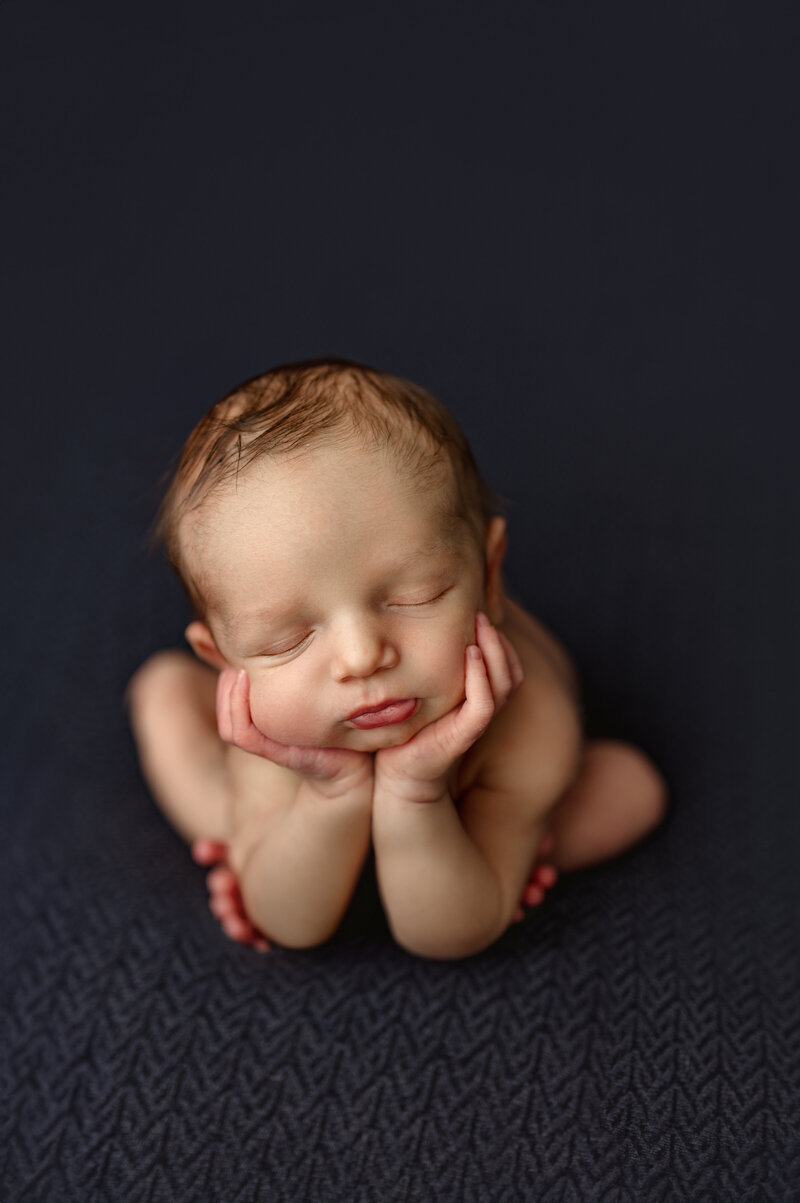 Newborn baby in a froggy pose resting peacefully on a textured dark blue blanket. Captured by Honeywild Photography, a Grand Rapids, MI, luxury newborn photographer specializing in timeless and elegant portraits