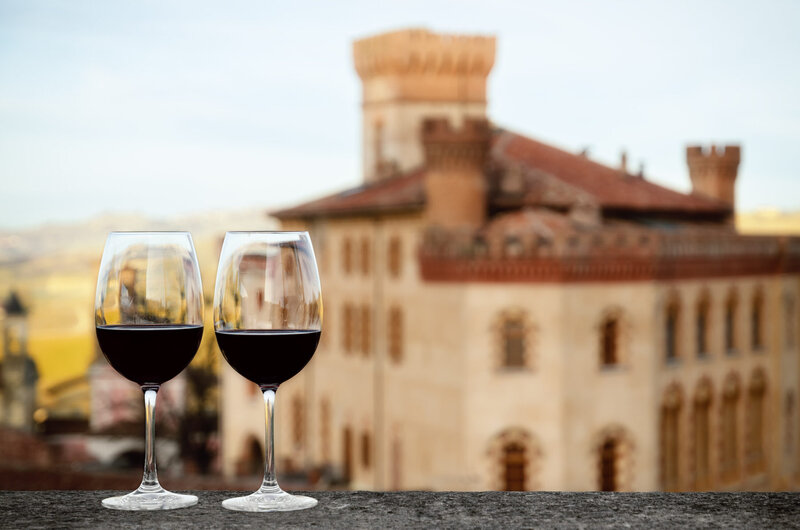 Two glasses of red wine sit on a ledge overlooking a blurred, historic castle with towers and red-tiled roofs in the background. The setting suggests a scenic view, possibly at sunset, perfect for those seeking luxury vacations with personalized travel planning.