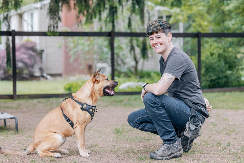 Dog Trainer Brittni Hyzer smiles at camera while medium sized brown dog looks at her obediently.
