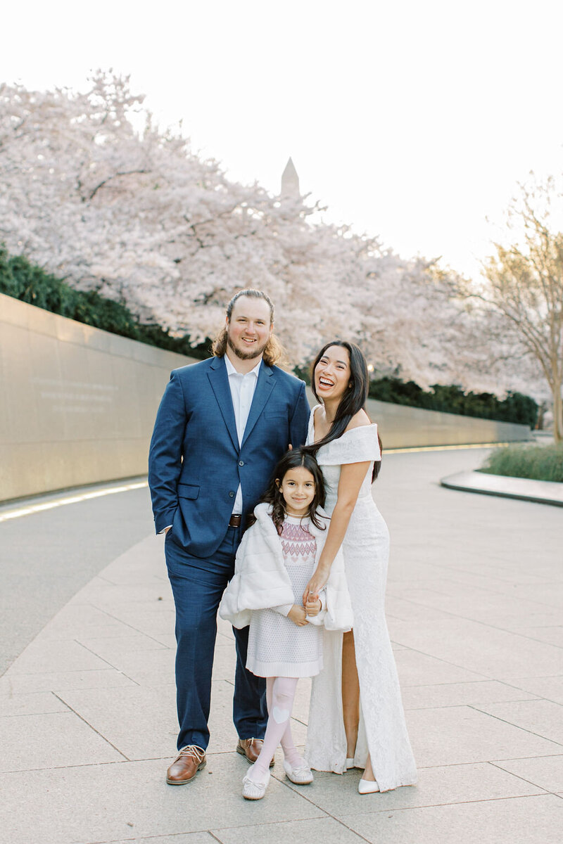 Husband, wife, and daughter smile together during cherry blossom season for family photos