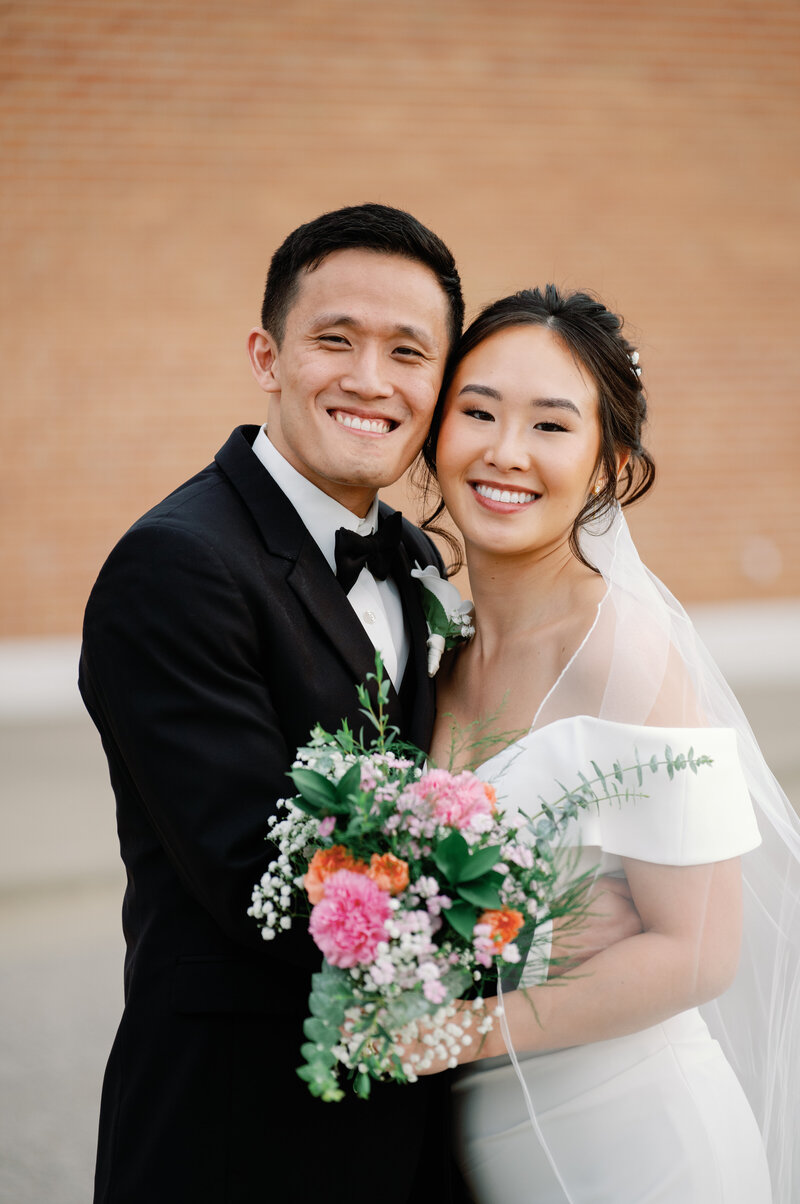 A Asian couple with a black tux and pink flowers smile for their wedding photo