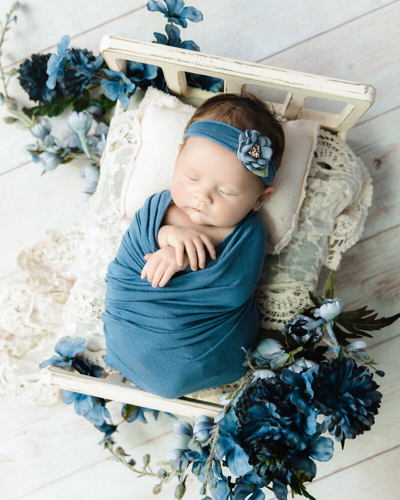 baby girl wrapped in blue sleeping on cream tiny bed with beautiful blue florals all around