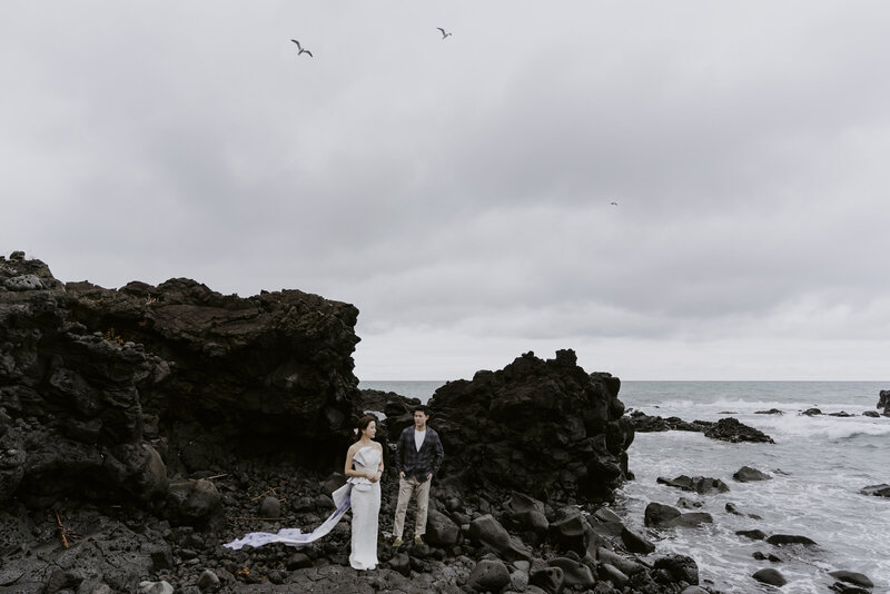 a bride in a wedding dress and a man in a suit stand on the coast in Jeju