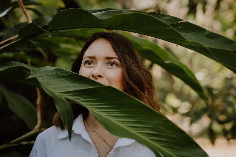 Brandi of The Intuitive Momma stands outdoors and peers over large, green leaves into distance.