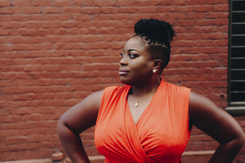 A black woman wearing a bright orange dress and braids pulled up on her head stands with her hands on her hips, looking confidently past the camera.