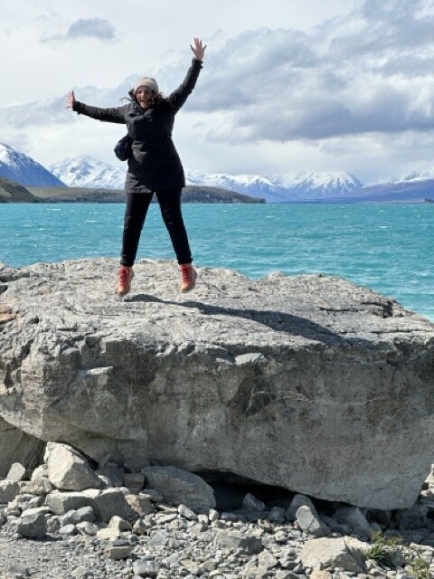 Maya jumps for joy on a rock in front of teal blue Lake Tekapo, New Zealand