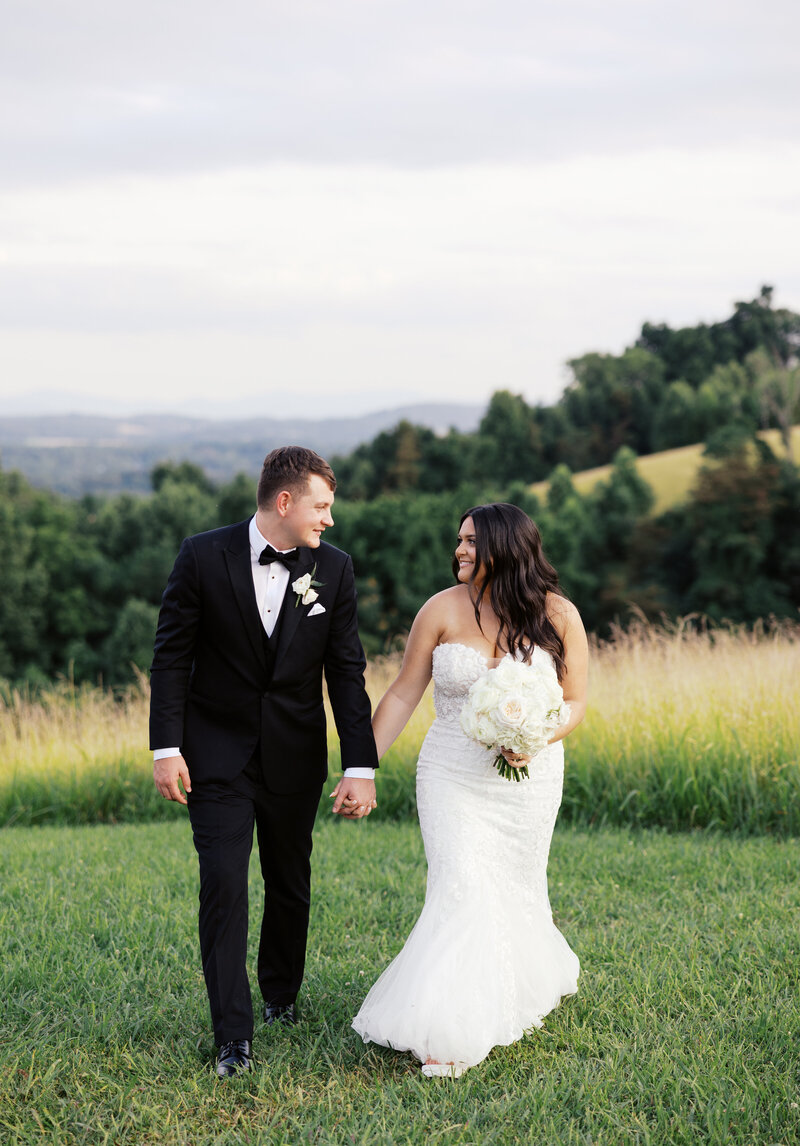 Couple walking together while looking at each other and smiling during sunset photos with mountains in the background.