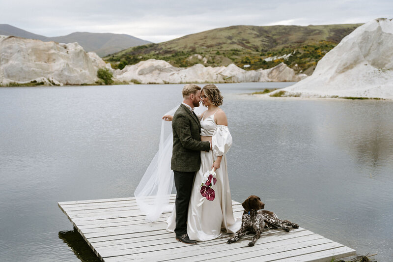 Bride and groom on a wooden dock with their dog, overlooking the lake at St Bathans in Central Otago.