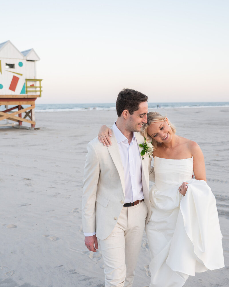 Photo of a bride and groom walking on the beach at sunset with a colorful building in the background