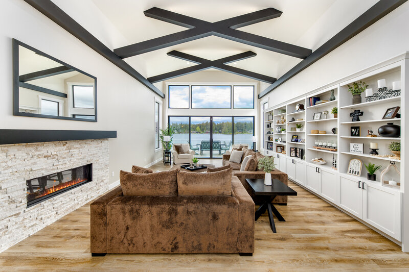 Contemporary living room with vaulted ceiling, exposed black beams, built-in white cabinetry, and lakefront views—featuring warm wood flooring and modern furnishings designed by Belle Design Collective.