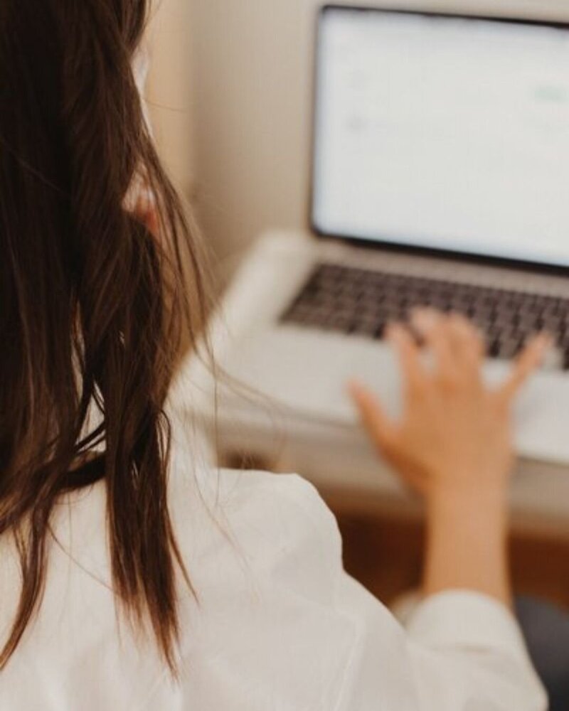A soft-focus image of a woman with long brown hair using a laptop at a simple desk.