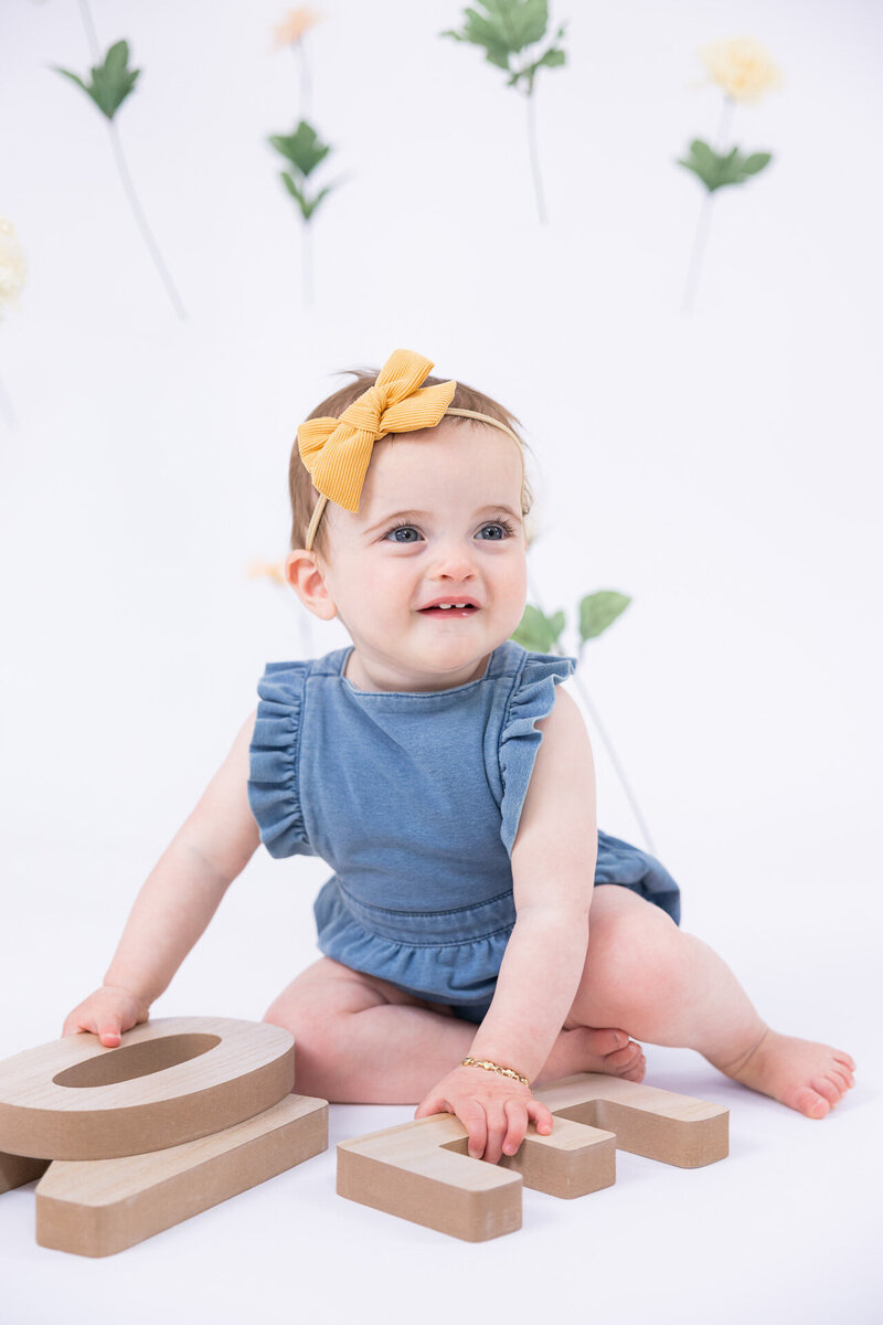 A cheerful baby girl in a blue ruffled romper and yellow bow headband sits on a white studio floor, smiling and playing with large wooden letters.