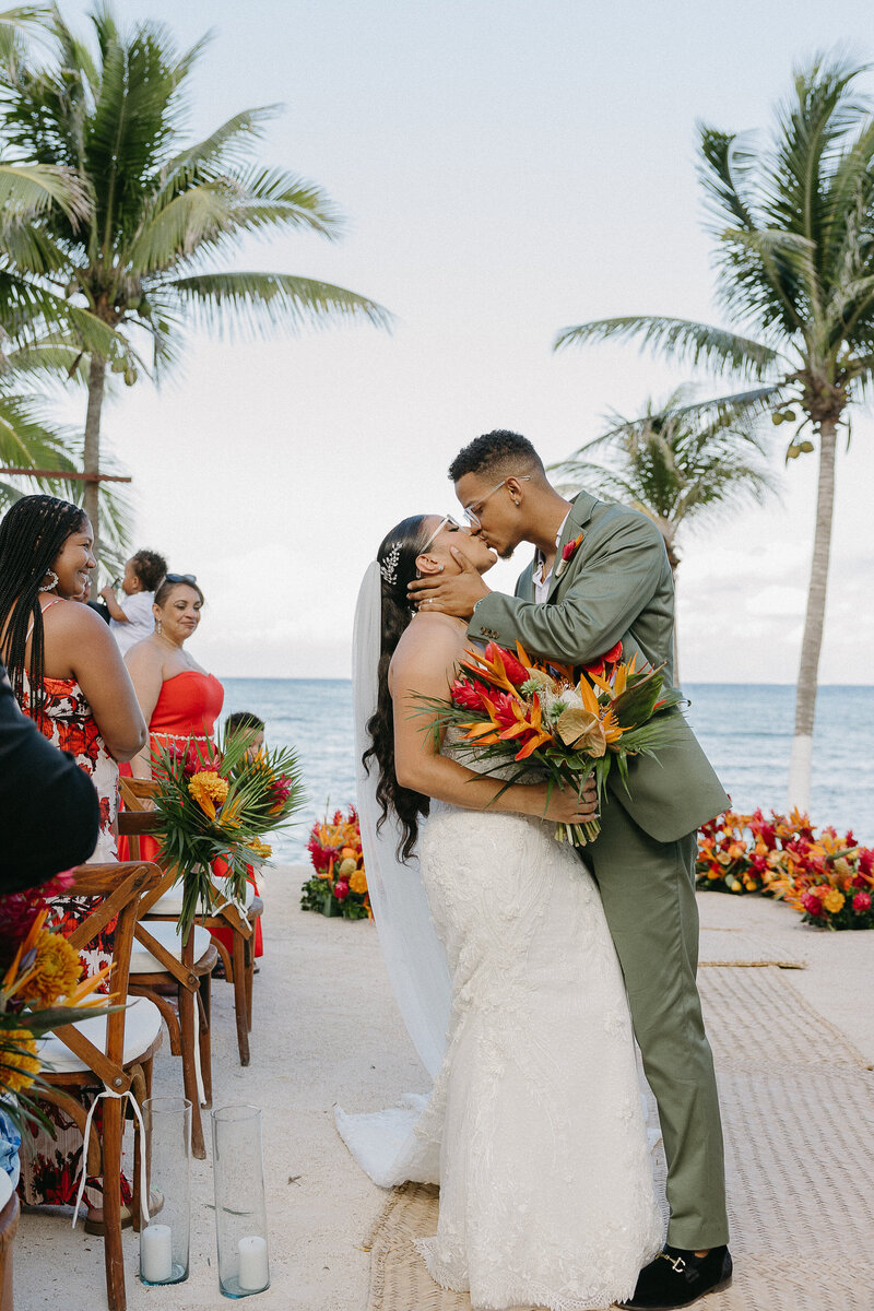 A newlywed couple shares a romantic kiss at their tropical beach wedding, surrounded by palm trees and vibrant floral arrangements in a Mexico destination wedding.
