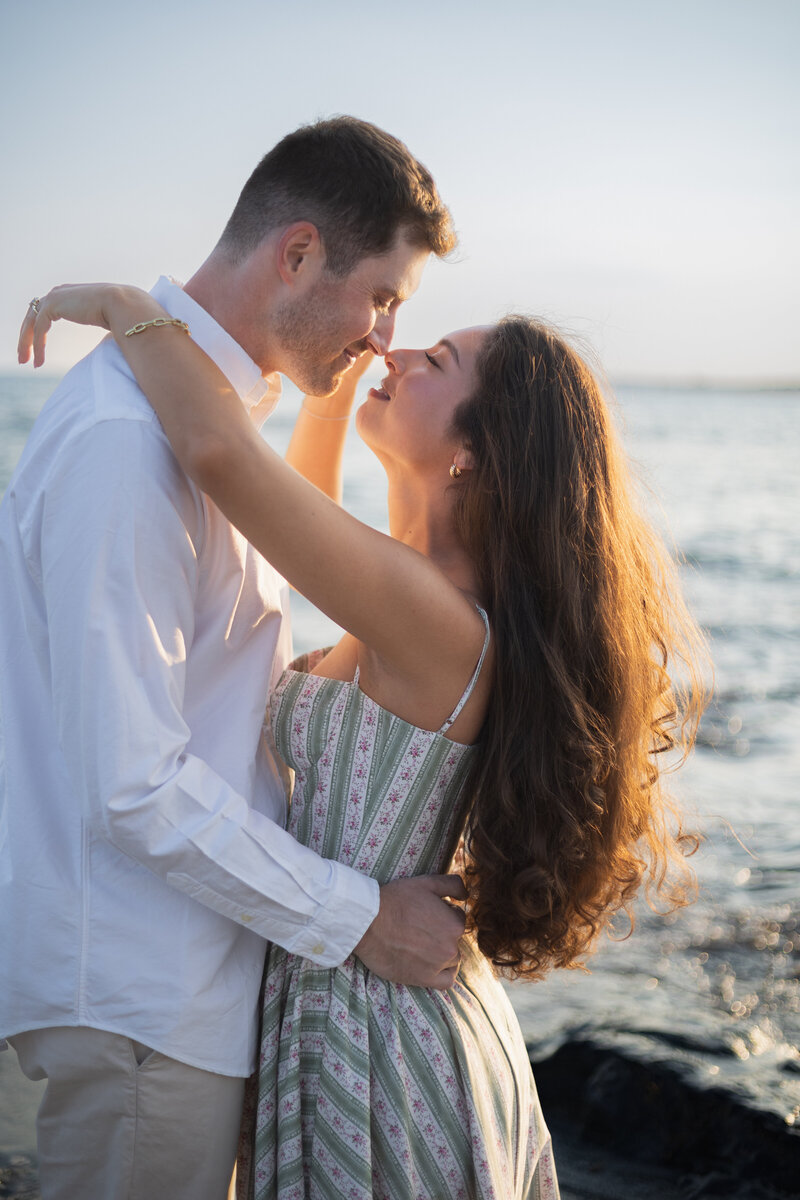 Collins Beach Newport RI | Kelsey Sheehan Photography Timeless Rhode Island Weddings | A couple embraces on a beach, smiling affectionately at each other. Sunset light highlights her flowing hair. Waves gently break in the background. Romantic, serene.