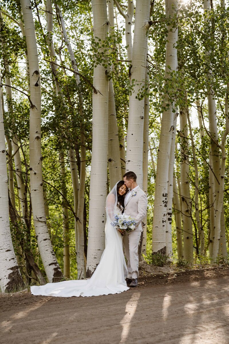 Bride and groom standing in a grove of aspen trees with soft sunlight and mountain forest scenery, photographed by a Colorado Wedding and Portrait Photographer.