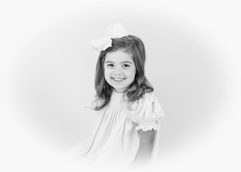 Black-and-white heirloom portrait of a smiling girl in a smocked dress with a bow in her hair.