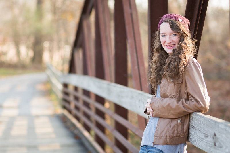 high school senior girl has her back leaning into a taller metal bridge with her elbows resting on a railing, she is wearing a brown leather jacket and a burgandy winter type toboggan, photographed by jamie lynette photography, canton ohio senior photographer