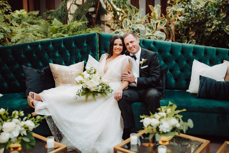Bride and groom smiling at the camera posed on a green couch surrounded by floral arrangements