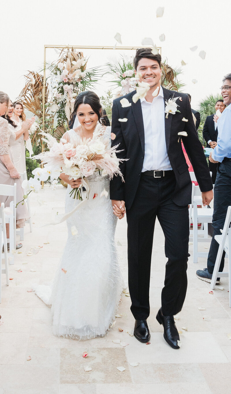 Bride and groom walk down the aisle with flowers and petals in a joyful outdoor wedding