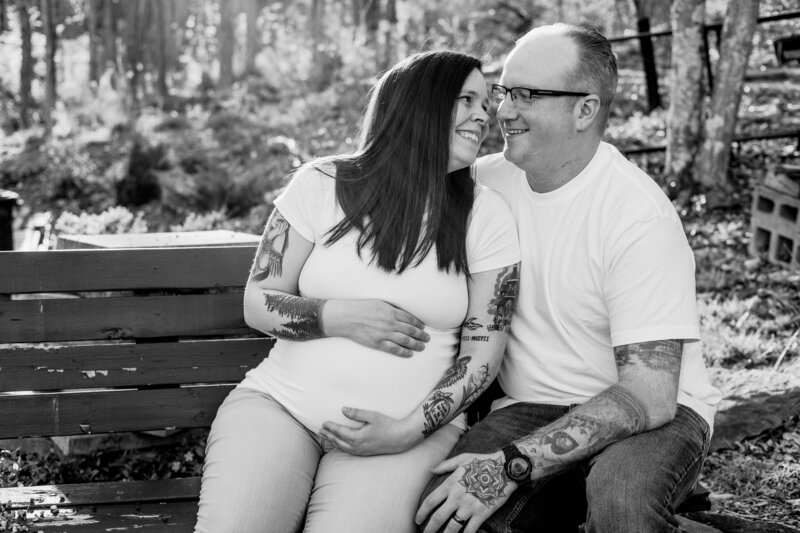 Black and white image of a  pregnant mom and dad sitting on a bench smiling at each other.