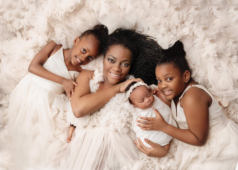 Greater Toronto Photographer studio of mom and her 3 daughters laying in white gowns holding baby girl at their studio session.