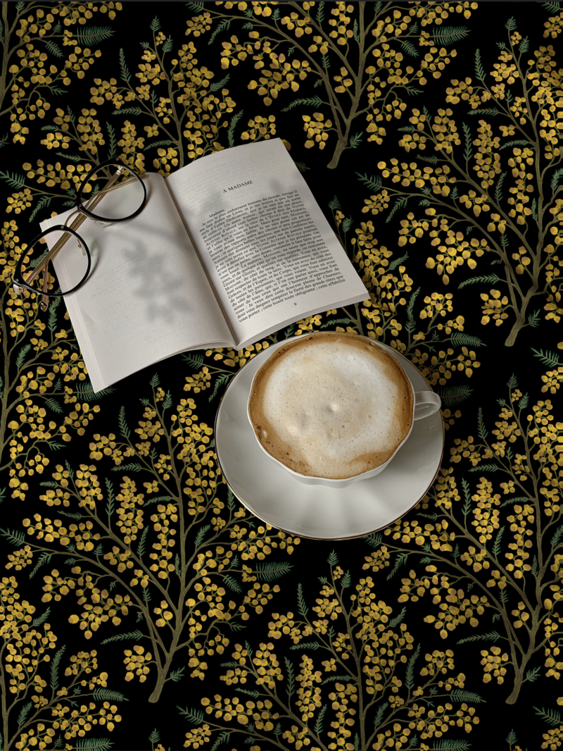 Floriography fabric tablecloth printed with vibrant hand drawn mimosa flowers is shown with an open book, reading glasses, and a frothy cappuccino. Fabric pattern is dark and moody, vintage in mustard yellow, sage green, black. National Women’s Day Flower in Italy