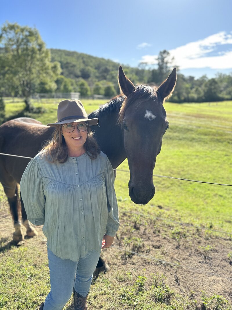 A young boy in a blue top, black pants and a cargo print hat brushing a dark brown horse
