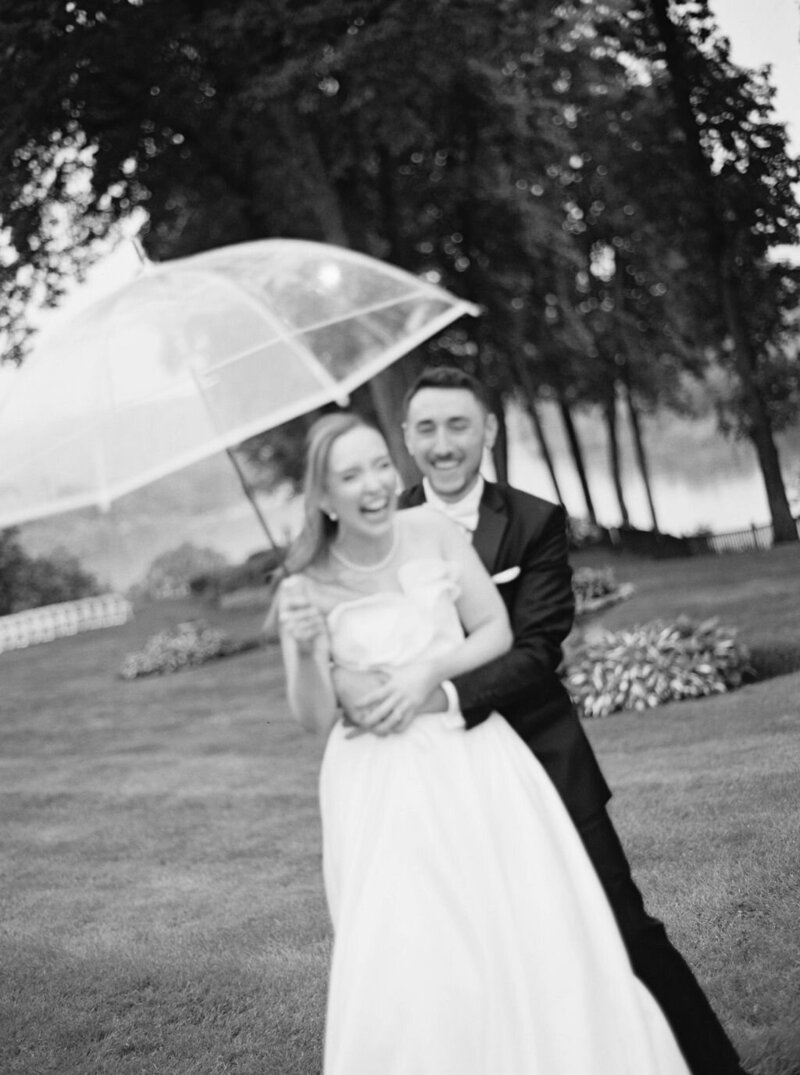 Rhode Island Wedding Photographers | A joyful couple in wedding attire laughs under a transparent umbrella in a lush garden. The black-and-white photo conveys a timeless, romantic mood.