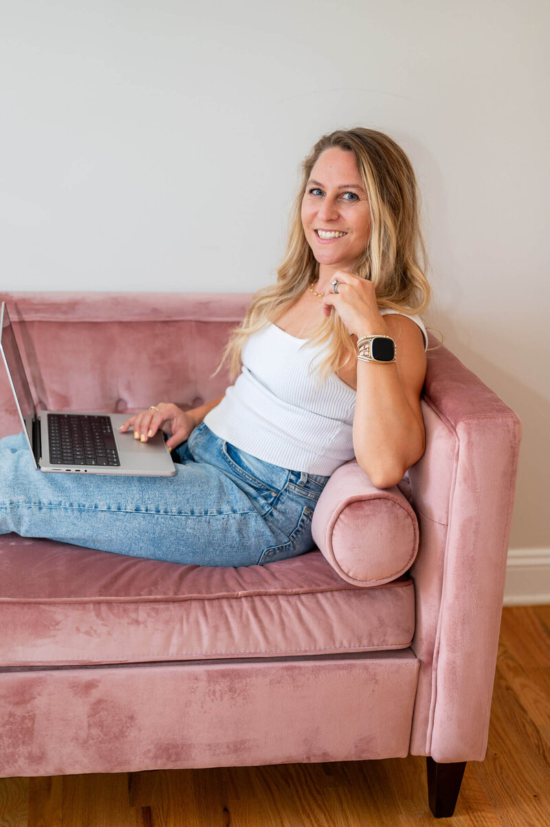 Marketing consultant sitting leaning back on pink velvet couch while working on her laptop, smiling at the camera in Westport, CT.