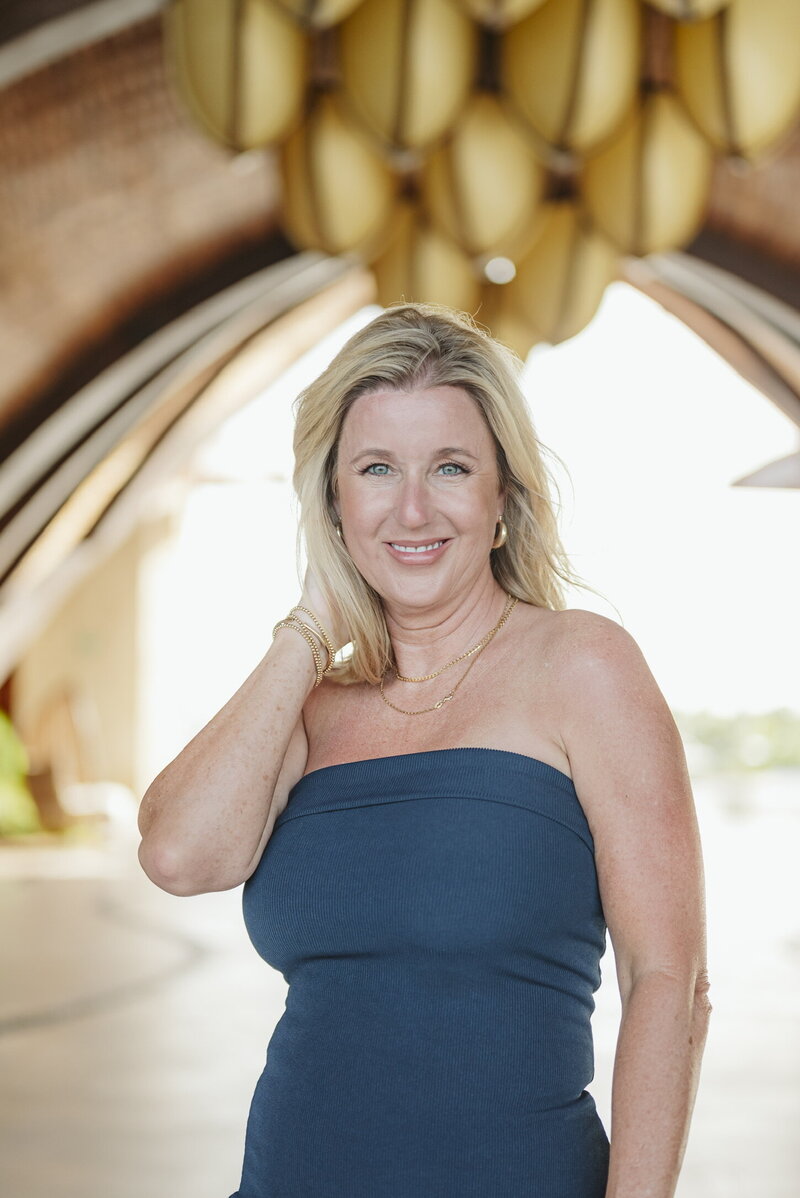 A portrait of a destination wedding planner wearing a strapless navy blue dress, standing outdoors under an architectural structure with soft natural lighting. She smiles while touching her hair.