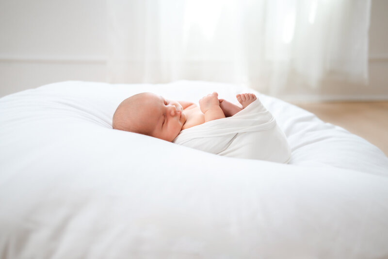 Baby boy wrapped in white swaddle sleeps on white beanbag.