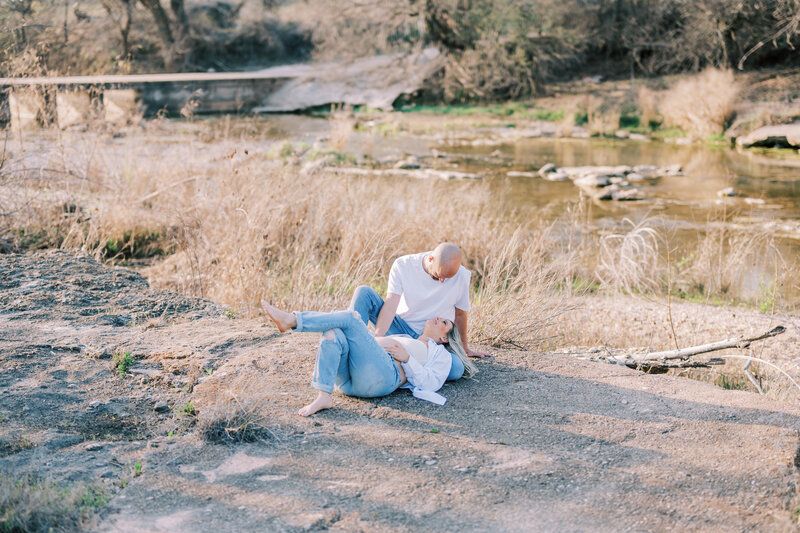 Woman laying on the ground with her head in man's lap in the grass in front of water