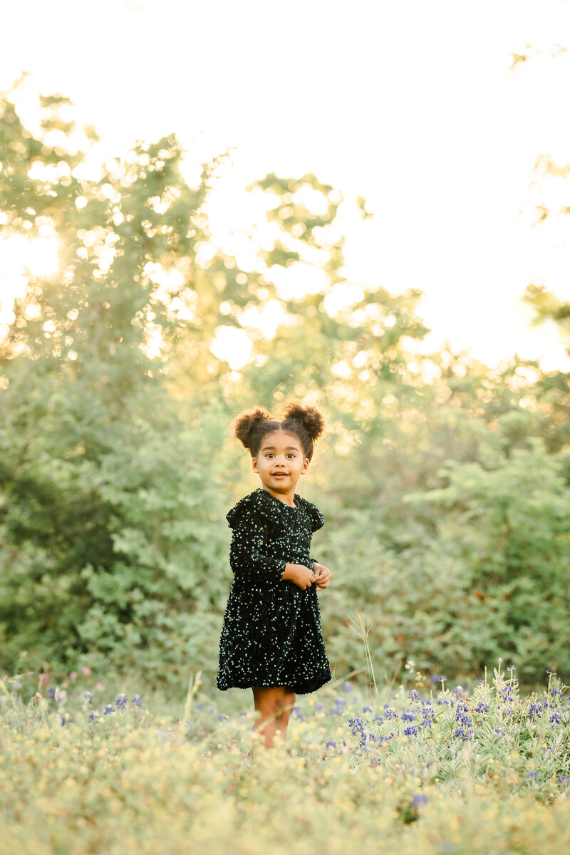 The photograph captures a serene and picturesque scene. A young girl, dressed in a dark dress, stands amidst a sprawling field of vibrant bluebonnets. Her gaze is directed straight at the camera, her expression radiating a warm and genuine smile.  The background is a stunning display of nature's beauty - a lush forest in the distance, its foliage ablaze with the vibrant hues of spring. The setting sun casts a warm, golden glow over the entire scene, creating a soft and ethereal atmosphere.  The photographer, Bri Sullivan, has masterfully utilized a light and airy style, allowing the natural beauty of the environment to take center stage. The image is well-balanced, with the girl's placement within the frame complementing the surrounding landscape perfectly.  The overall effect is one of tranquility and joy, inviting the viewer to pause and immerse themselves in the serene moment captured by the camera. The girl's radiant smile and the stunning natural backdrop work in harmony, creating a truly captivating and memorable photograph.