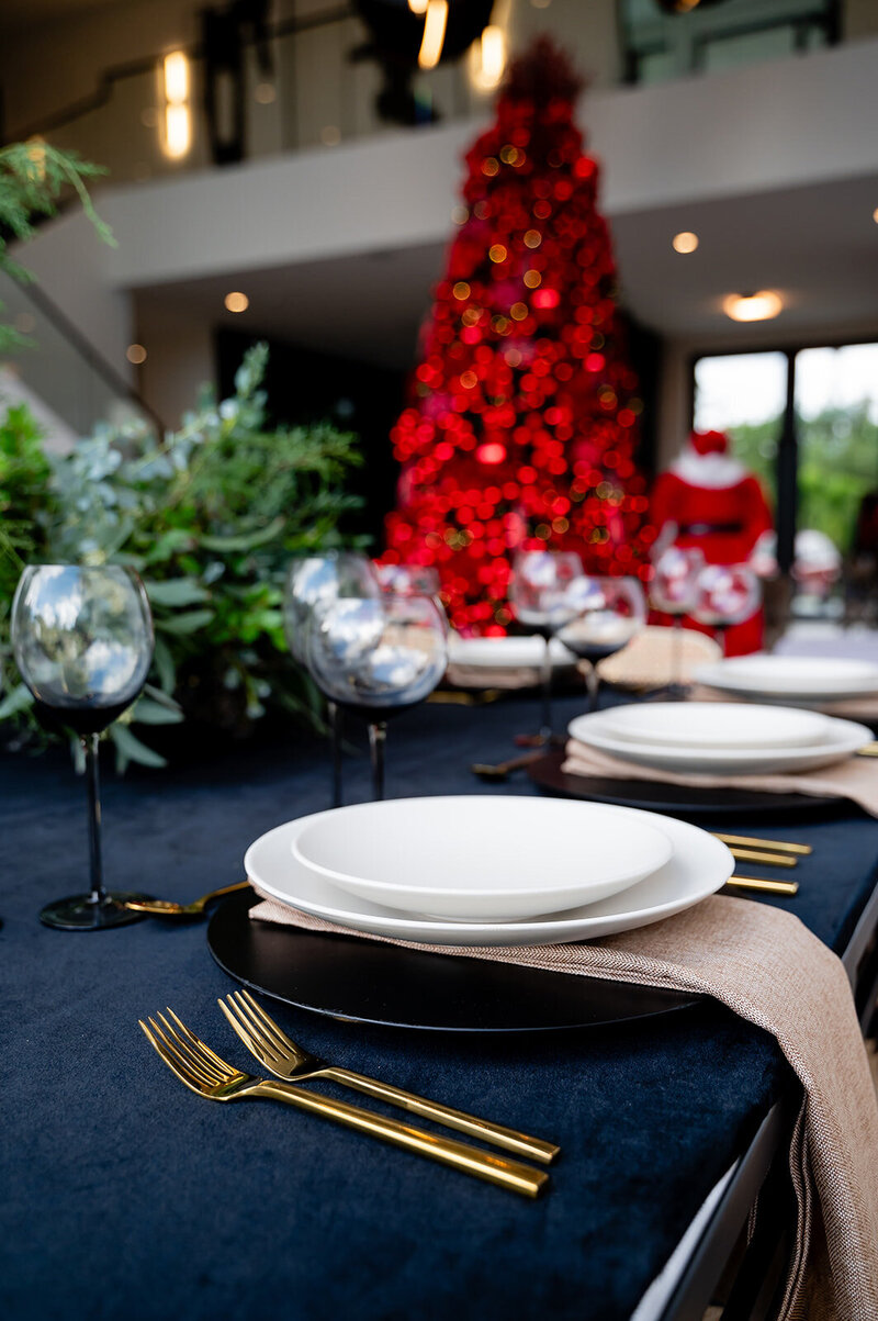 Elegant holiday dining table with black tablecloth, glassware, and neutral place settings in front of a large red Christmas tree, showcasing the Luxe Holiday Experience by Interiors by CV.