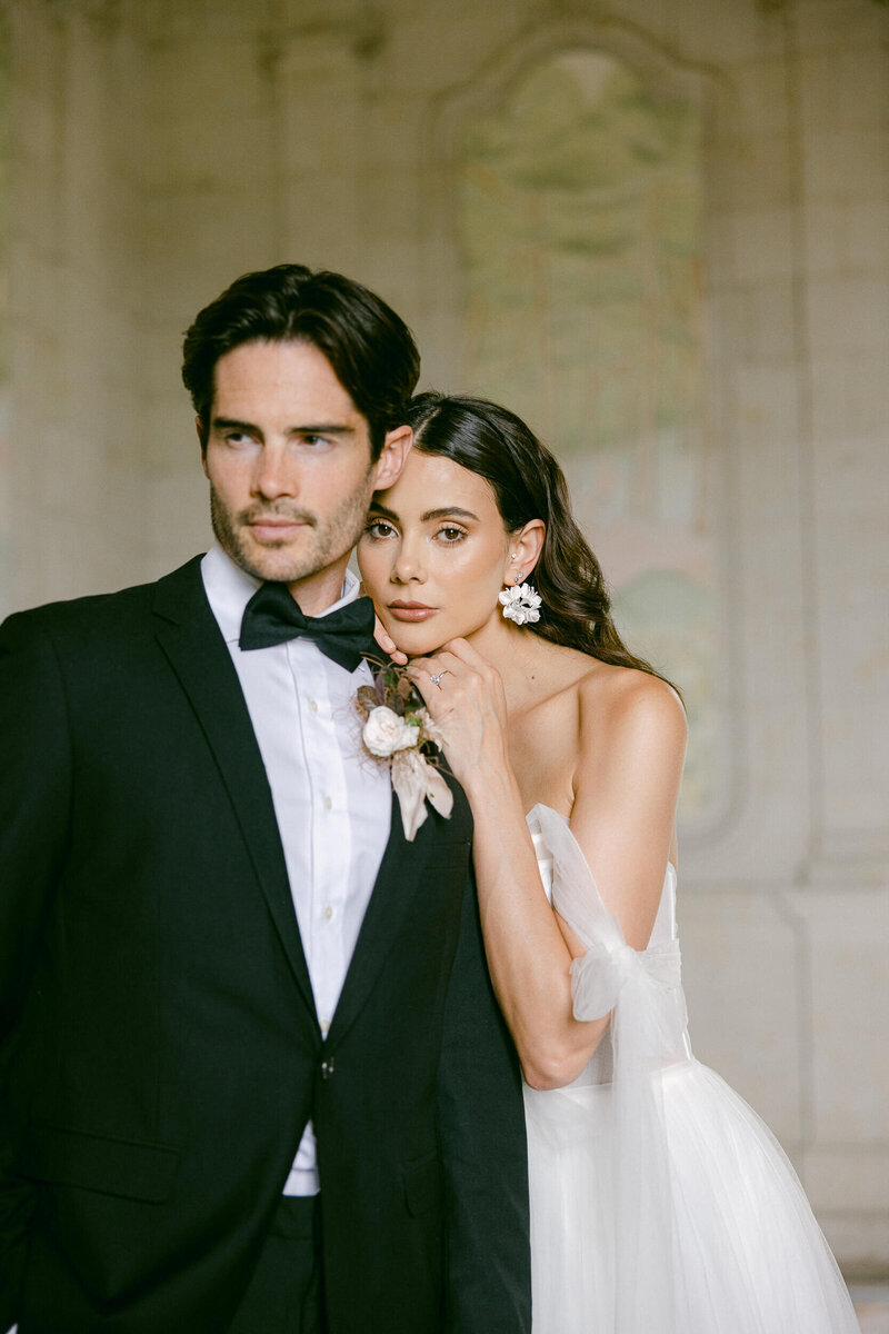 a picture of a bride and groom with the bride standing behind the groom with her head and hands resting on the grooms shoulder as they pose for a photo on their wedding day