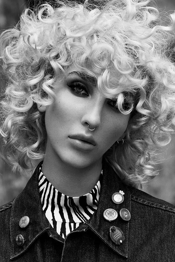 Platinum blonde curls styled into a voluminous shape, paired with a denim jacket adorned with vintage pins and a striped scarf.