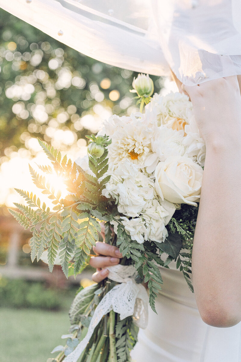 Bride holding a cream and white bouquet with roses, ferns, and lace ribbon in golden sunset light.