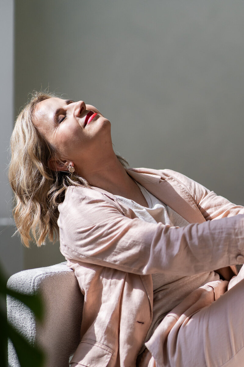 žena ve světlém kostýmku sedí na křesle, má zavřené oči a hlavu zakloněnou ke slunci